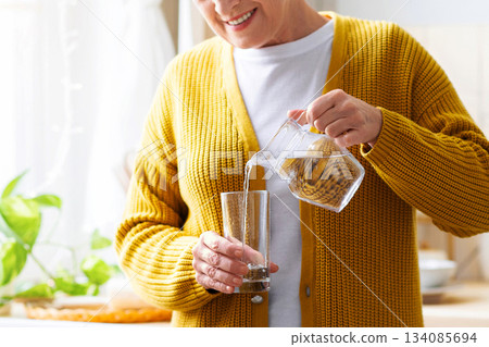 Hydration in senior ages concept. Unrecognizable grandmother standing in cozy white kitchen, pouring water into glass. Cropped of senior woman having healthy lifestyle, drinking water at home Hydration in senior ages concept. Unrecognizable grandmother standing in cozy white kitchen, pouring water into glass. Cropped of senior woman having healthy lifestyle, drinking water at home 134085694