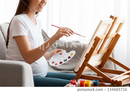 Pregnancy Leisure. Happy pregnant woman painting at home, sitting on couch near easel, holding color palette, smiling expectant mother enjoying drawing art therapy, side view with copy space Pregnancy Leisure. Happy pregnant woman painting at home, sitting on couch near easel, holding color palette, smiling expectant mother enjoying drawing art therapy, side view with copy space 134085710
