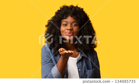 Emotional beautiful african american yougn lady with booshie hairstyle sending flying kiss to camera over yellow studio background, copy space. Love, emotions, feelings concept 134085735