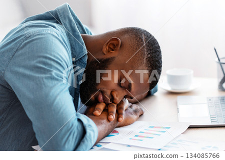 Tired african american freelancer guy sleeping at desk with laptop and papers, young black man napping at workplace in home office, exhausted male placed head on hands, tired after long work 134085766