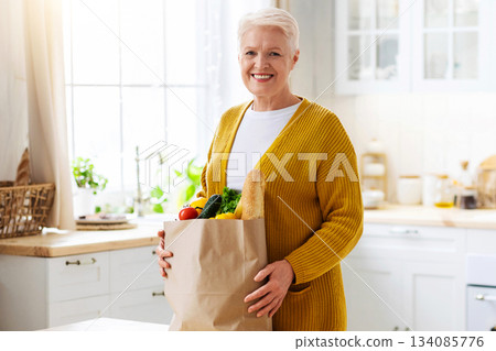 Smiling old woman checking paper bag with grocery ordered from internet during COVID-19 pandemic, kitchen interior, copy space. Cheerful senior lady unpacking purchase after shopping. Food delivery Smiling old woman checking paper bag with grocery ordered from internet during COVID-19 pandemic, kitchen interior, copy space. Cheerful senior lady unpacking purchase after shopping. Food delivery 134085776