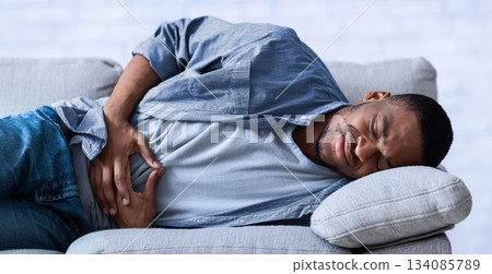 Stomachache. African Guy Suffering From Abdominal Pain Touching Aching Stomach Lying On Sofa At Home. Black Man Having Gastritis Problem And Abdomen Inflammation. Health Issue Concept Stomachache. African Guy Suffering From Abdominal Pain Touching Aching Stomach Lying On Sofa At Home. Black Man Having Gastritis Problem And Abdomen Inflammation. Health Issue Concept 134085789