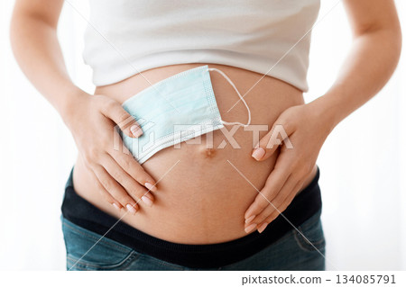Pregnancy And Pandemic Concept. Closeup Of Pregnant Woman Holding Protective Medical Mask Near Belly, Standing Near Window, Expectant Lady Keeping Preventive Measures Against Coronavirus Outbreak Pregnancy And Pandemic Concept. Closeup Of Pregnant Woman Holding Protective Medical Mask Near Belly, Standing Near Window, Expectant Lady Keeping Preventive Measures Against Coronavirus Outbreak 134085791