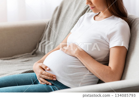 Prenatal Time. Beautiful Future Mother Sitting On Couch Embracing Her Belly, Happy Pregnant Woman Relaxing on Sofa In Living Room At Home, Tenderly Touching Tummy And Smiling, Side View, Free Space Prenatal Time. Beautiful Future Mother Sitting On Couch Embracing Her Belly, Happy Pregnant Woman Relaxing on Sofa In Living Room At Home, Tenderly Touching Tummy And Smiling, Side View, Free Space 134085793
