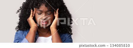 Closeup of upset african american young lady in jeans coat with curly hair having headache, touching her temples, closing eyes, grey studio background. Migraine, headache, tension, cluster concept 134085810