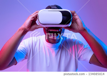 Virtual Gaming Concept. Portraif Of Funny Black Guy Wearing VR Glasses And Exclaiming With Excitement While Playing Video Game, Standing In Neon Light Over Purple Studio Background, Closeup Virtual Gaming Concept. Portraif Of Funny Black Guy Wearing VR Glasses And Exclaiming With Excitement While Playing Video Game, Standing In Neon Light Over Purple Studio Background, Closeup 134085814