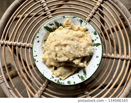 Tempe Mendoan, an Indonesian traditional snack made from tempe or tempeh (fermented soybeans) coated in flour and spring onions and fried half-cooked, on a white enamel plate, placed on a rattan table Tempe Mendoan, an Indonesian traditional snack made from tempe or tempeh (fermented soybeans) coated in flour and spring onions and fried half-cooked, on a white enamel plate, placed on a rattan table 134086165