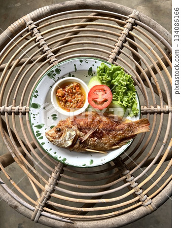 Fried tilapia served on a white enamel plate with a green rim, accompanied by chili sauce, and fresh vegetables specifically lettuce, sliced cucumber, and tomato. Placed on a rattan woven table. 134086166