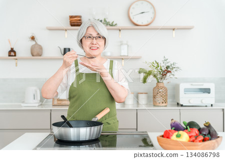一位身穿圍裙的老婦人在家裡的廚房裡一邊做飯一邊品嚐食物。 一位身穿圍裙的老婦人在家裡的廚房裡一邊做飯一邊品嚐食物。 134086798
