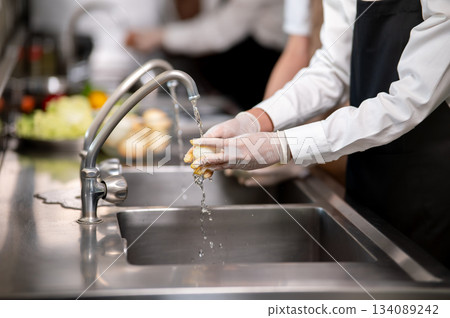 Close up of gloved hands chef washing or cleaning a peeled onion with tab water in kitchen sink. Close up of gloved hands chef washing or cleaning a peeled onion with tab water in kitchen sink. 134089242