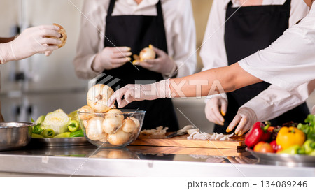 Close up of gloved hands chef and students or assistant peeling onions together at kitchen counter. Close up of gloved hands chef and students or assistant peeling onions together at kitchen counter. 134089246