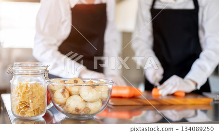 Bowl of onions and jar of fettuccine or ribbon pasta with blurred background of hands cutting carrot Bowl of onions and jar of fettuccine or ribbon pasta with blurred background of hands cutting carrot 134089250