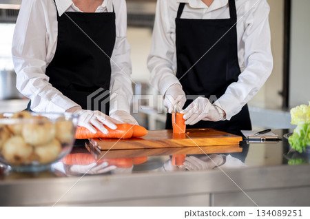 Close up of hands in rubber gloves holding knife cutting carrot on chopping board at kitchen counter Close up of hands in rubber gloves holding knife cutting carrot on chopping board at kitchen counter 134089251