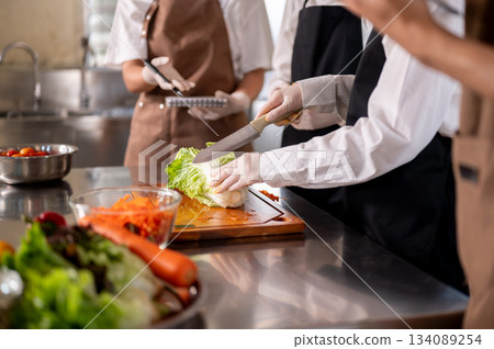 Chef holding knife cutting cabbage on chopping board with students or assistants at kitchen counter Chef holding knife cutting cabbage on chopping board with students or assistants at kitchen counter 134089254