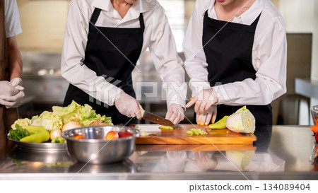 Female chef looking and teaching an assistant to cut a pepper on chopping board at kitchen counter. 134089404