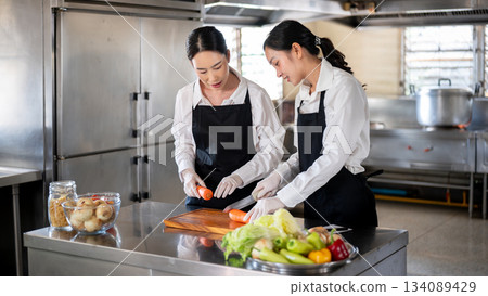 Student or assistant cutting carrot with knife while female chef teaching at kitchen cooking counter Student or assistant cutting carrot with knife while female chef teaching at kitchen cooking counter 134089429