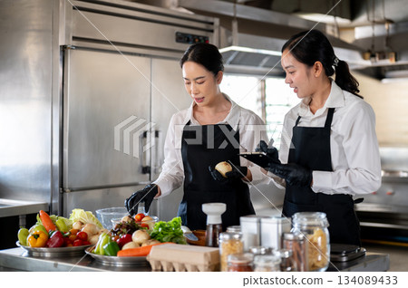 Student or assistant note down while female chef teaching at kitchen counter with food ingredients. 134089433