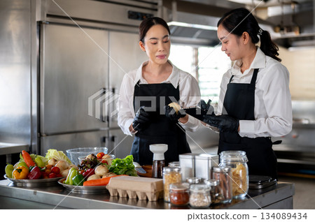Female chef teaching a student or assistant in kitchen at cooking counter full of food ingredients. Female chef teaching a student or assistant in kitchen at cooking counter full of food ingredients. 134089434