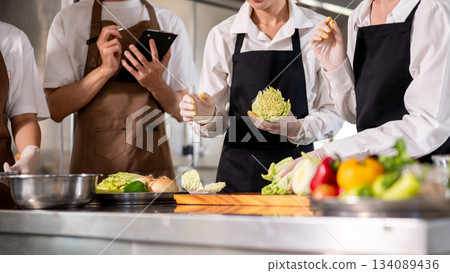 Close up of chef holding cabbage cuts while standing with students or assistants at kitchen counter. Close up of chef holding cabbage cuts while standing with students or assistants at kitchen counter. 134089436