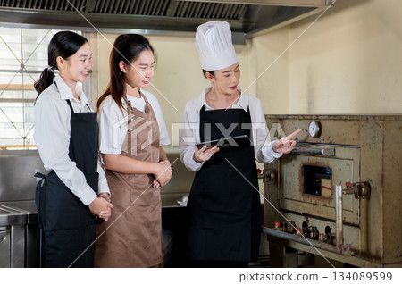 Asian female chef holding a tablet and pointing at oven thermometer to her students in the kitchen. Asian female chef holding a tablet and pointing at oven thermometer to her students in the kitchen. 134089599