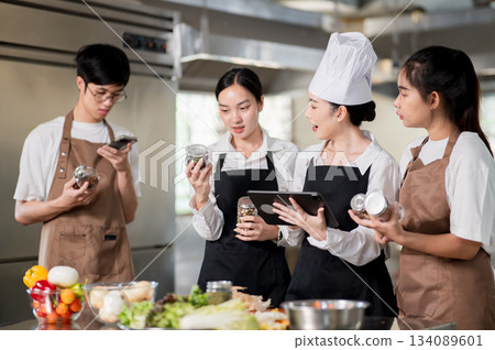 Asian female chef holding a tablet while her students looking at seasonal jars over kitchen counter. Asian female chef holding a tablet while her students looking at seasonal jars over kitchen counter. 134089601