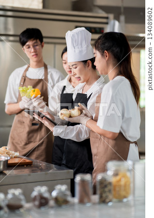 Asian female chef holding a tablet reading recipe to students or assistants over a kitchen counter. 134089602