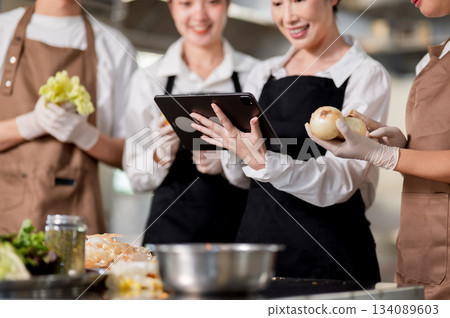 Asian female chef holding a tablet showing recipe to her students or assistants over kitchen counter 134089603