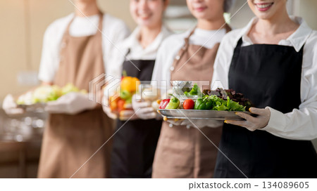 Close up of chefs with gloves and apron holding stainless plate and glass bowl with fresh vegetables Close up of chefs with gloves and apron holding stainless plate and glass bowl with fresh vegetables 134089605