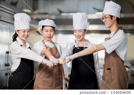 Asian male and female chefs wearing gloves and apron standing and making a fist bump in the kitchen. Asian male and female chefs wearing gloves and apron standing and making a fist bump in the kitchen. 134089606