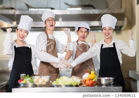 Asian chefs wearing gloves with apron are joining hands and raising their fist over a kitchen counter Asian chefs wearing gloves with apron are joining hands and raising their fist over a kitchen counter 134089611
