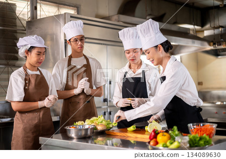 Female chef picking bell peppers while her students or assistants standing around the kitchen counter. Female chef picking bell peppers while her students or assistants standing around the kitchen counter. 134089630