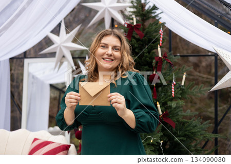 Young 30s woman getting Christmas greeting card from her beloved on background of xmas tree at cozy living room with festive interior. Surprised lady pleasure reading letter at home. 134090080