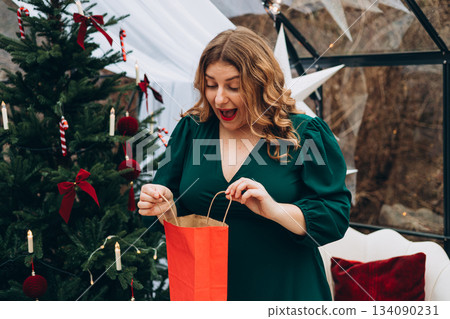 Middle age Caucasian woman in dress smiling confident holding Christmas gift bag at home. Winter time and holidays. Celebration banner, big sale concept 134090231