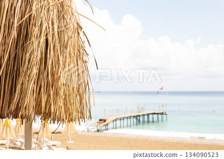 Thatched roof with tropical beach with sand and wooden pier on the sea shore in background. Thatched roof with tropical beach with sand and wooden pier on the sea shore in background. 134090523