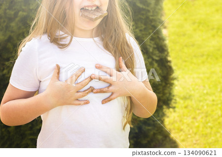 Girl child staining her white t-shirt with stain while enjoying a delicious chocolate bar in garden 134090621