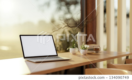 White screen laptop and a coffee cup on wooden counter in a glass wall cafe with nature view outside White screen laptop and a coffee cup on wooden counter in a glass wall cafe with nature view outside 134090749