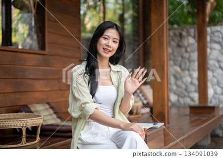 Long hair asian woman smiling and waving her hand while sitting cross legged at wooden house terrace 134091039