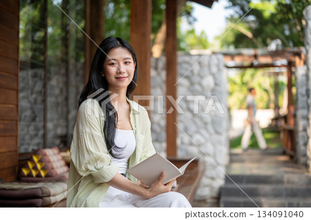 Long hair asian woman smiling while holding white cover book sitting cross legged at wooden terrace 134091040