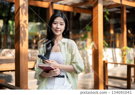 Asian woman holding books and smartphone while sitting on railing or bench in garden wooden corridor Asian woman holding books and smartphone while sitting on railing or bench in garden wooden corridor 134091041