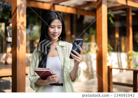 Asian woman is holding books and looking at her smartphone while sitting in garden wooden corridor. Asian woman is holding books and looking at her smartphone while sitting in garden wooden corridor. 134091042