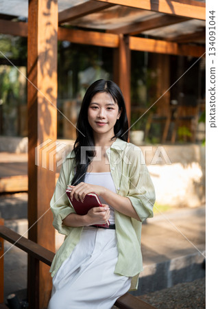 Long hair asian woman is holding books and sitting on a railing or a bench in garden wooden corridor. Long hair asian woman is holding books and sitting on a railing or a bench in garden wooden corridor. 134091044