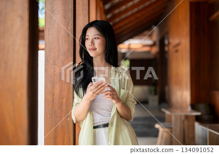 Long hair asian woman is leaning against a pillar while holding a phone in wooden house's corridor. Long hair asian woman is leaning against a pillar while holding a phone in wooden house's corridor. 134091052
