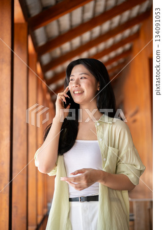 Long hair asian woman is talking on a smartphone and looking outside from a wooden house's corridor. Long hair asian woman is talking on a smartphone and looking outside from a wooden house's corridor. 134091053