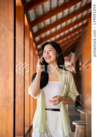 Long hair asian woman talking on a phone and looking ahead while walking in a wooden house's corridor. Long hair asian woman talking on a phone and looking ahead while walking in a wooden house's corridor. 134091054