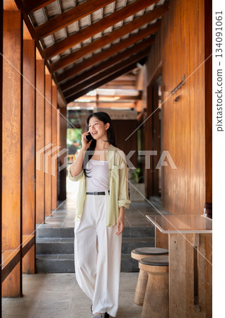 Long hair asian woman talking on a smartphone while standing or walking in wooden house's corridor. Long hair asian woman talking on a smartphone while standing or walking in wooden house's corridor. 134091061