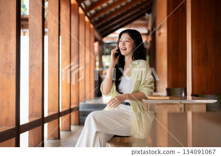 Long hair asian woman is talking on smartphone while sitting at table in wooden house's corridor. Long hair asian woman is talking on smartphone while sitting at table in wooden house's corridor. 134091067