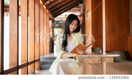 Long hair asian woman is reading a book while sitting at table in wooden house's corridor. Long hair asian woman is reading a book while sitting at table in wooden house's corridor. 134091068