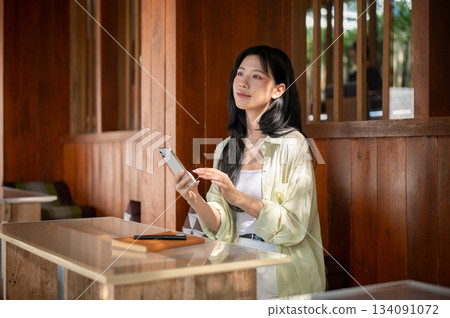 Long hair asian woman is looking away from her smartphone while sitting at table in a wooden house. Long hair asian woman is looking away from her smartphone while sitting at table in a wooden house. 134091072