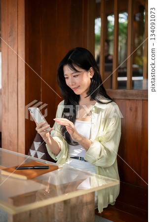 Asian woman smiling and looking at smartphone holding in hand while sitting at table in wooden house Asian woman smiling and looking at smartphone holding in hand while sitting at table in wooden house 134091073