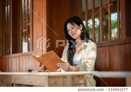 Long hair asian woman is smiling and reading a book while sitting at table in a wooden house. 134091077